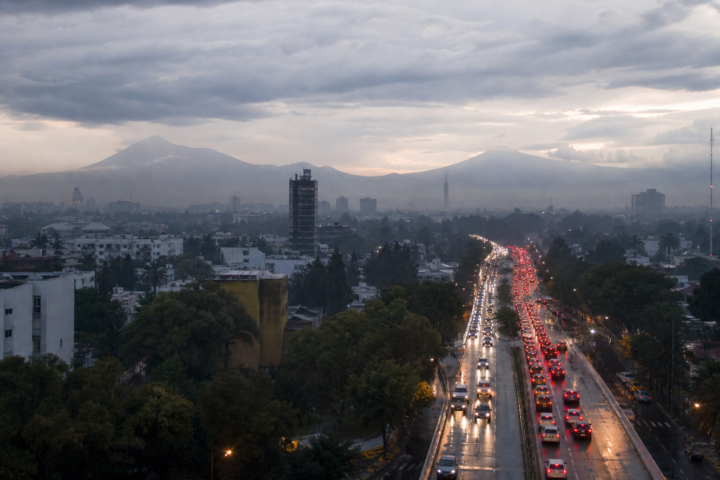 Tráfico vehicular en una avenida de la Ciudad de México al amanecer, con pavimento mojado y cielo nublado.