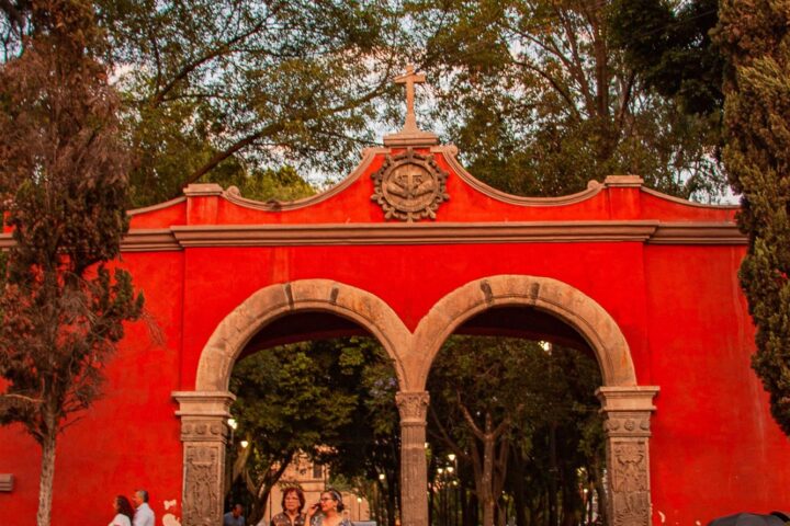 Imagen ilustrativa: acceso peatonal con arco y visitantes en un espacio público; no identifica la sede del evento.