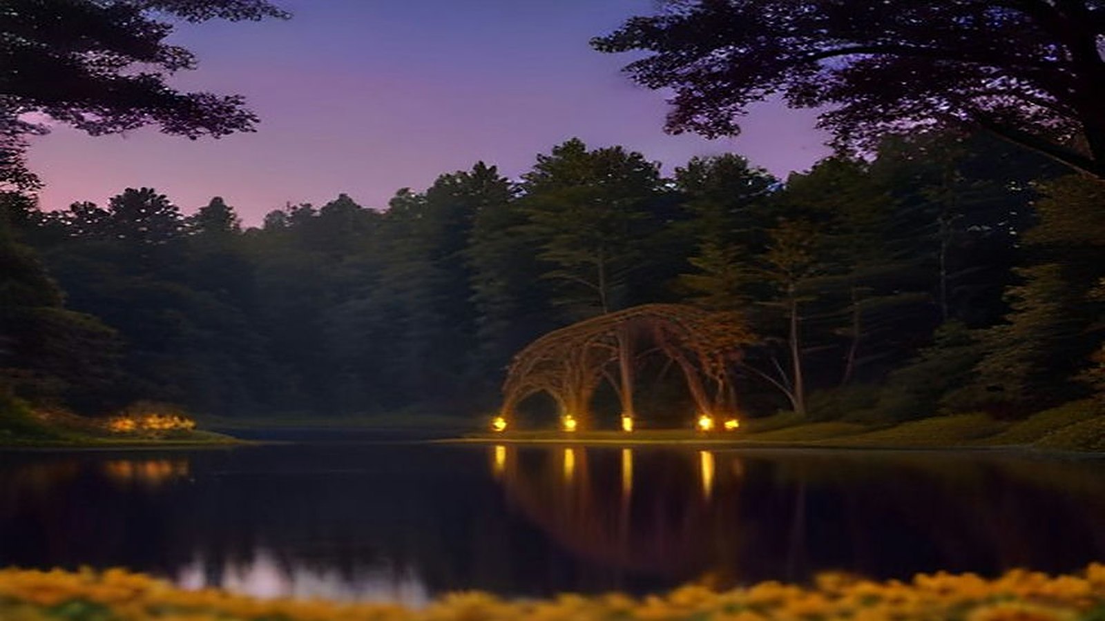 Lago de un bosque al anochecer con una estructura de arcos iluminada en la orilla, árboles altos, cielo morado y flores amarillas en primer plano
