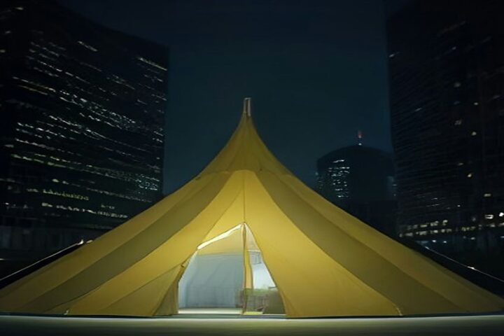 Gran carpa de circo amarilla iluminada de noche en una explanada, frente a rascacielos de vidrio con ventanas encendidas