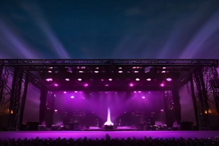 Escenario de festival de música al aire libre con estructura de torres de iluminación, bocinas y reflectores morados apuntando al cielo nocturno