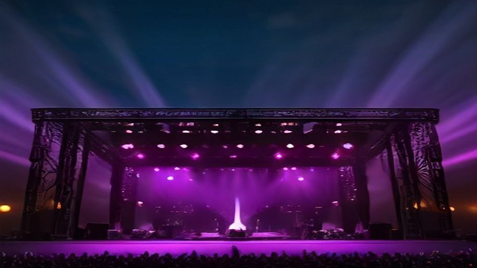 Escenario de festival de música al aire libre con estructura de torres de iluminación, bocinas y reflectores morados apuntando al cielo nocturno