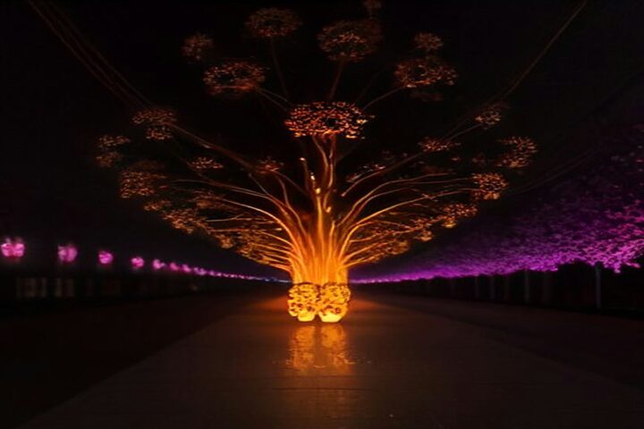 Escultura luminosa en forma de árbol con destellos anaranjados y una hilera de luces magenta sobre una avenida oscura de noche