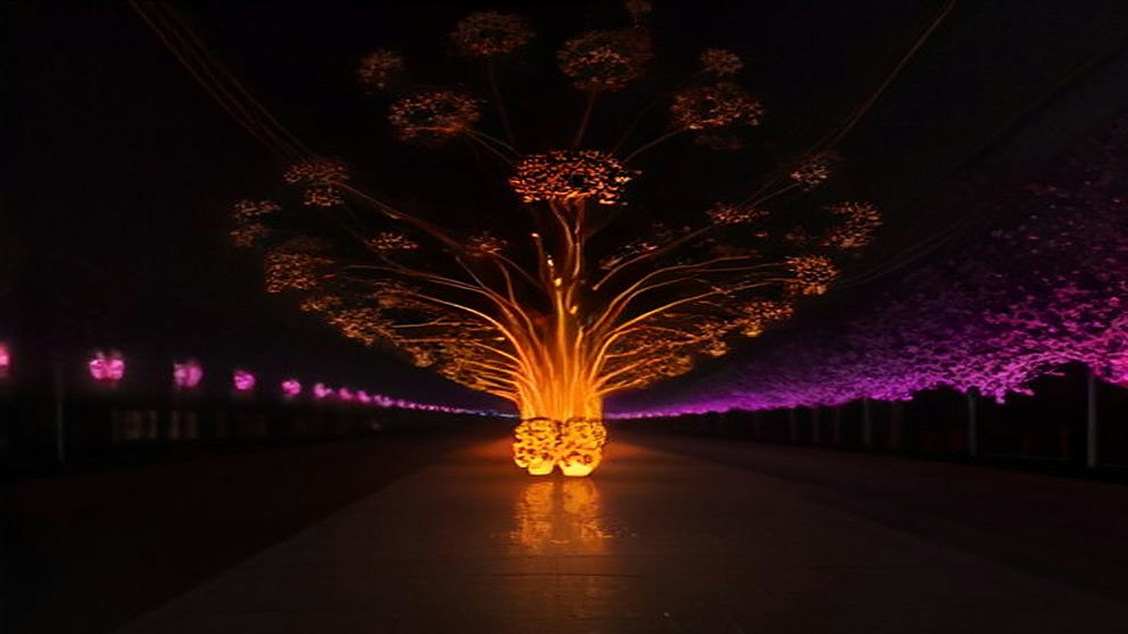 Escultura luminosa en forma de árbol con destellos anaranjados y una hilera de luces magenta sobre una avenida oscura de noche