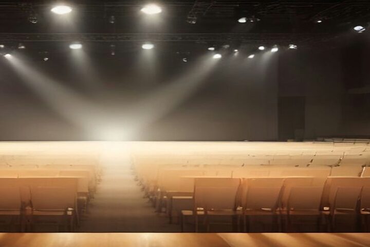 Escenario de concierto vacío con haces de luz blanca y cálida que atraviesan la bruma y largas filas de butacas sin público