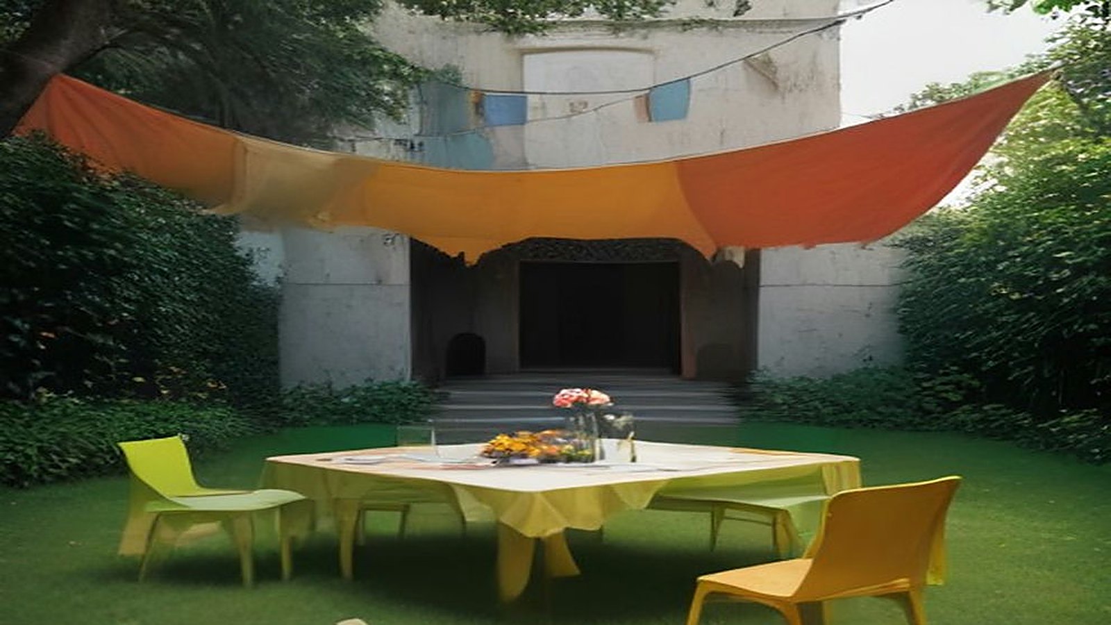 Jardín de un centro cultural con una lona anaranjada, banderines de colores, una mesa con flores de cempasúchil y sillas de colores sobre el pasto
