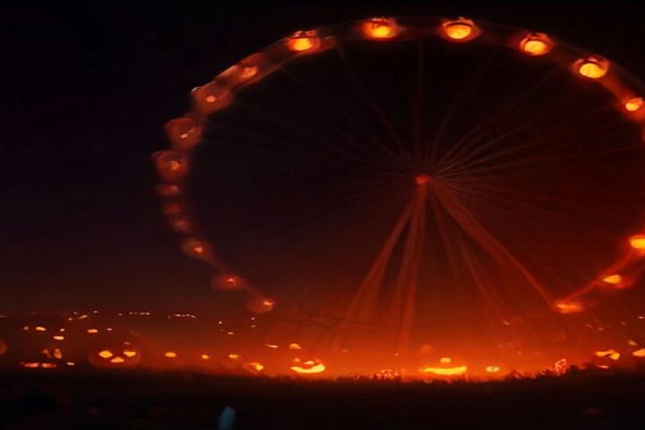 Rueda de la fortuna iluminada con luces naranjas en medio de la neblina nocturna, con decenas de calabazas encendidas sobre el pasto de un parque vacío