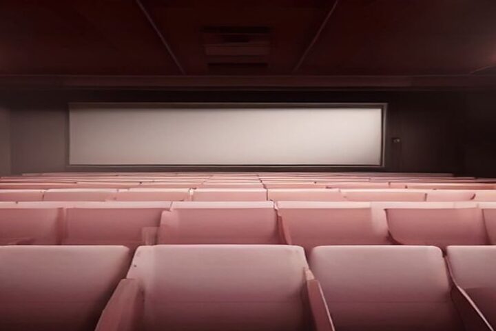 Sala de cine vacía con filas de butacas color rosa frente a una gran pantalla blanca encendida, en penumbra