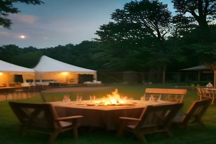 Mesa de madera con una fogata encendida y copas en un jardín al atardecer, con carpas blancas iluminadas al fondo