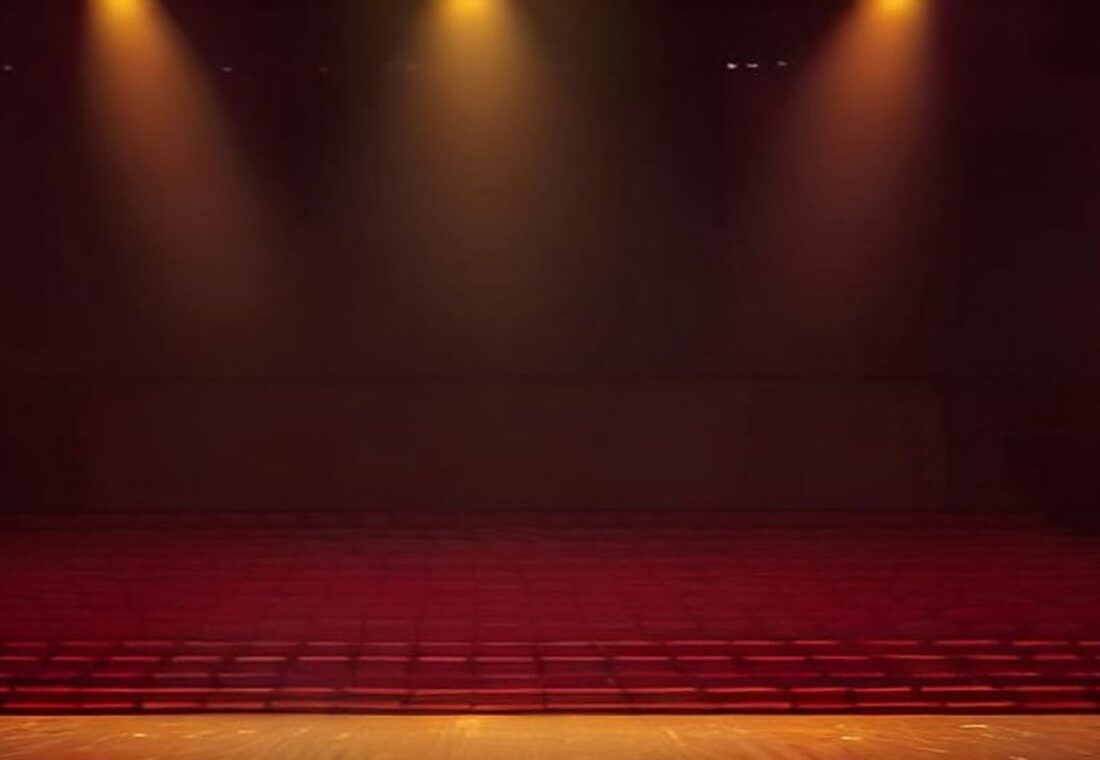Escenario de un gran auditorio vacío iluminado por reflectores dorados, con destellos en el aire y filas de butacas rojas en penumbra