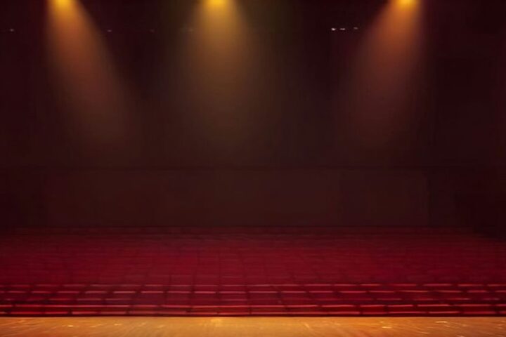 Escenario de un gran auditorio vacío iluminado por reflectores dorados, con destellos en el aire y filas de butacas rojas en penumbra
