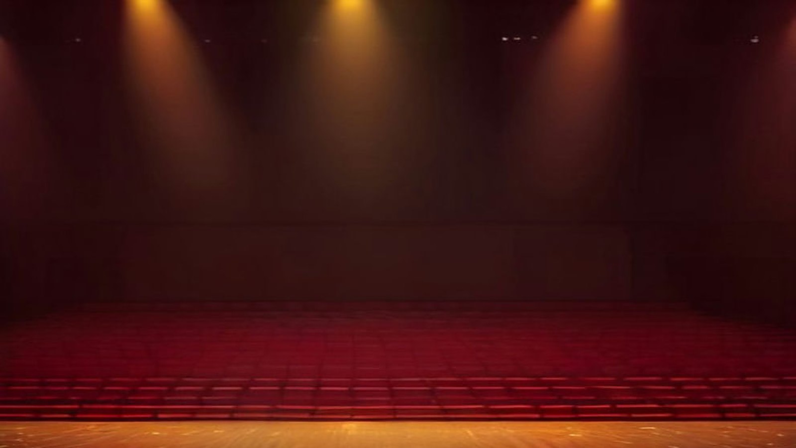 Escenario de un gran auditorio vacío iluminado por reflectores dorados, con destellos en el aire y filas de butacas rojas en penumbra