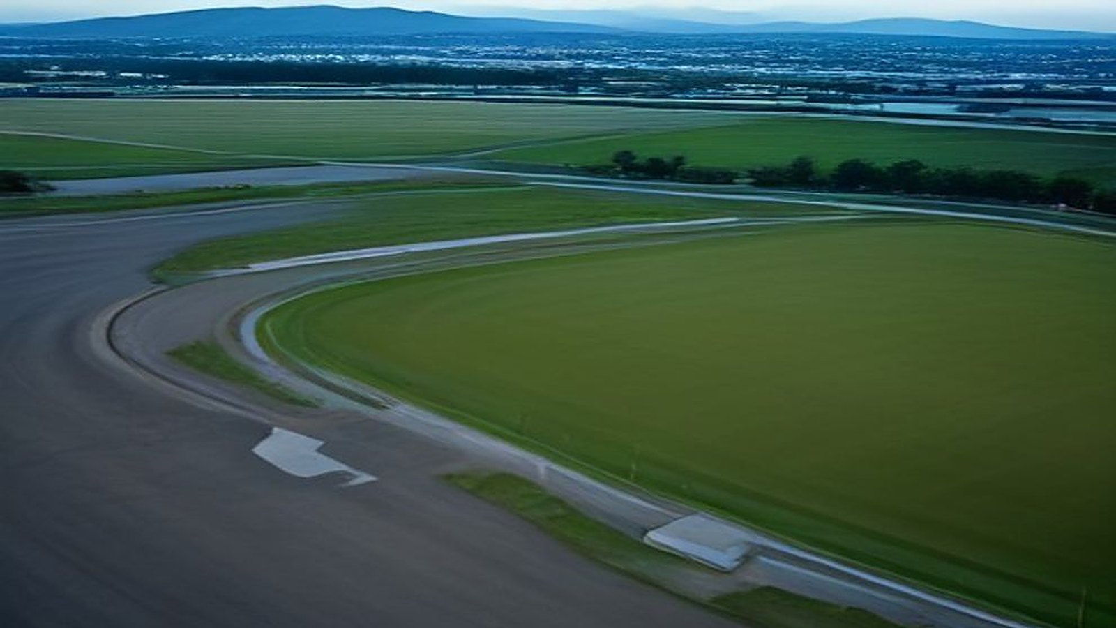 Vista aérea de un circuito de carreras vacío al atardecer, con curvas de asfalto, zonas de pasto verde y la ciudad con montañas al fondo