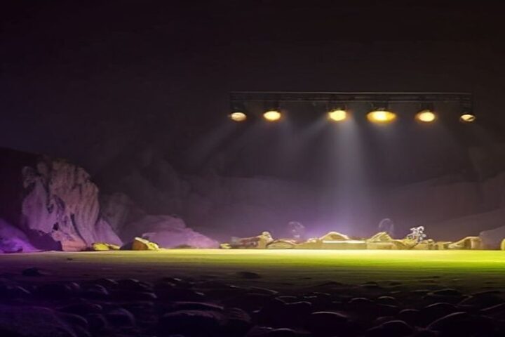 Escenario de festival vacío dentro de una cantera de roca por la noche, con luces moradas, verdes y anaranjadas que iluminan la neblina y las paredes de piedra