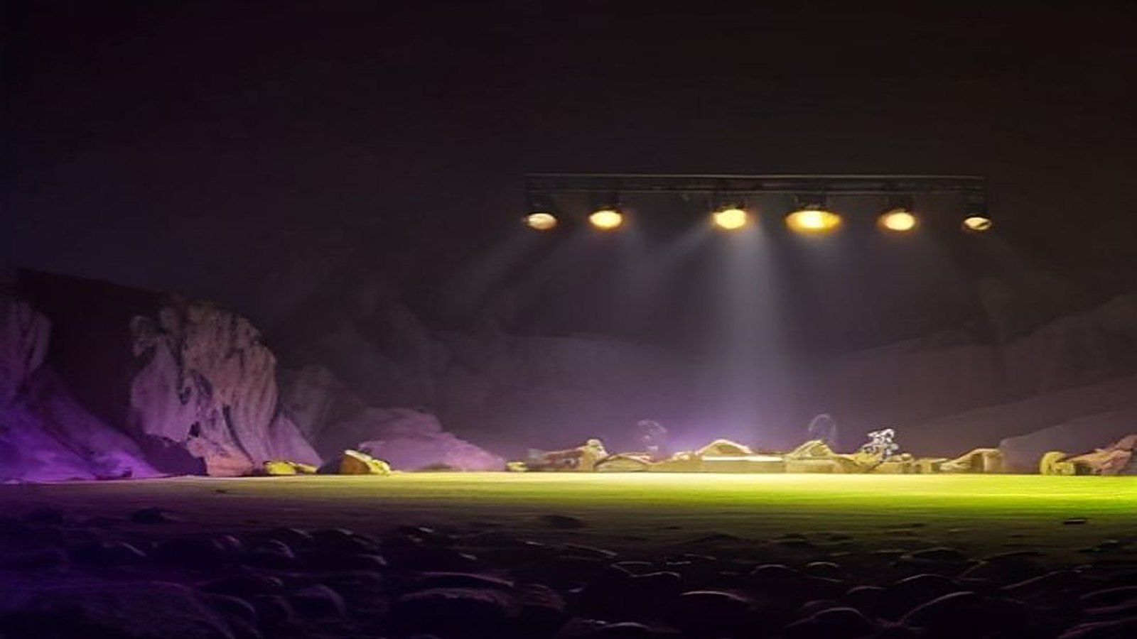 Escenario de festival vacío dentro de una cantera de roca por la noche, con luces moradas, verdes y anaranjadas que iluminan la neblina y las paredes de piedra