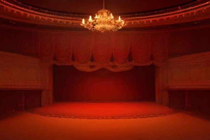 Interior de un teatro antiguo con candelabro, cortinas rojas y el escenario vacío iluminado en tonos rojos y naranjas