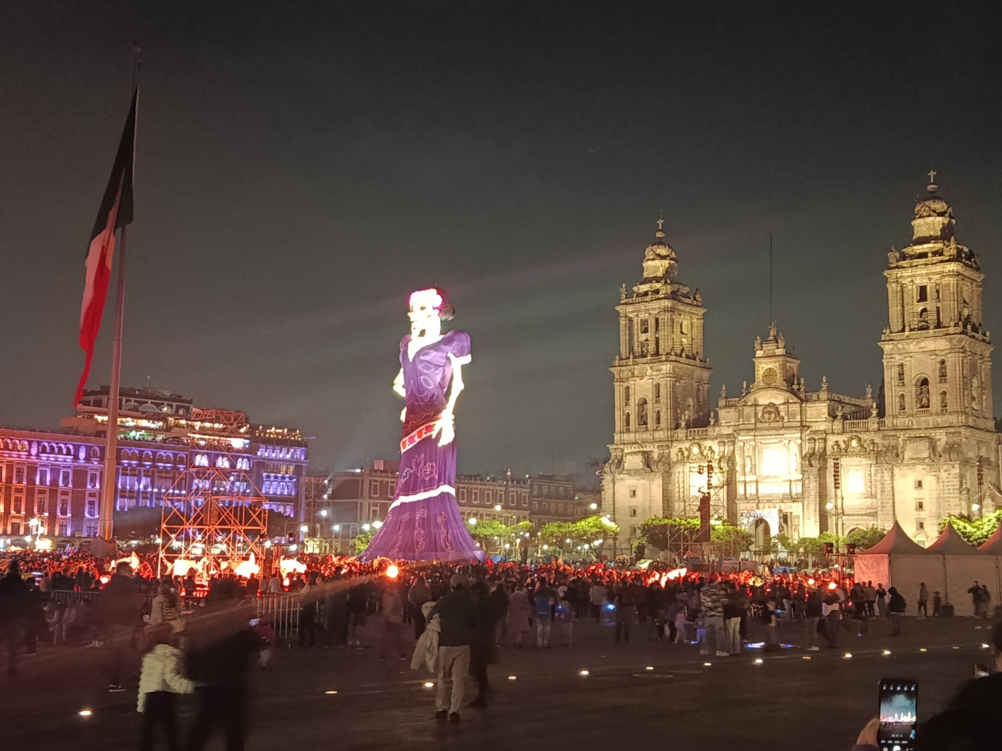 Catrina monumental iluminada en el Zócalo de la Ciudad de México, junto a la Catedral Metropolitana, de noche y con visitantes