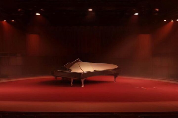 Escenario de sala de conciertos con un piano de cola bajo luces rojas y doradas, alfombra roja y butacas vacías en penumbra