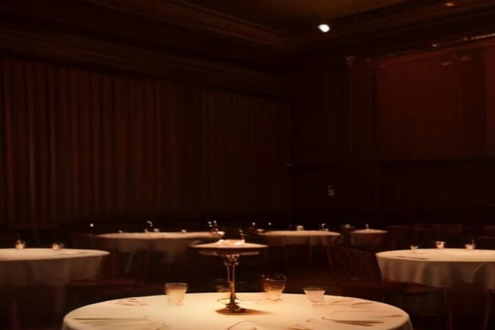 Salón de cabaret en penumbra con mesas redondas de mantel blanco, copas y una lámpara de mesa bajo luz cálida