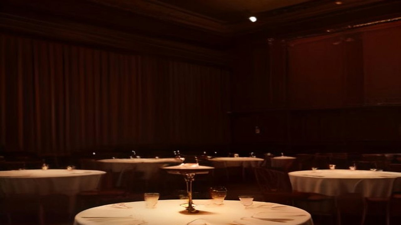 Salón de cabaret en penumbra con mesas redondas de mantel blanco, copas y una lámpara de mesa bajo luz cálida