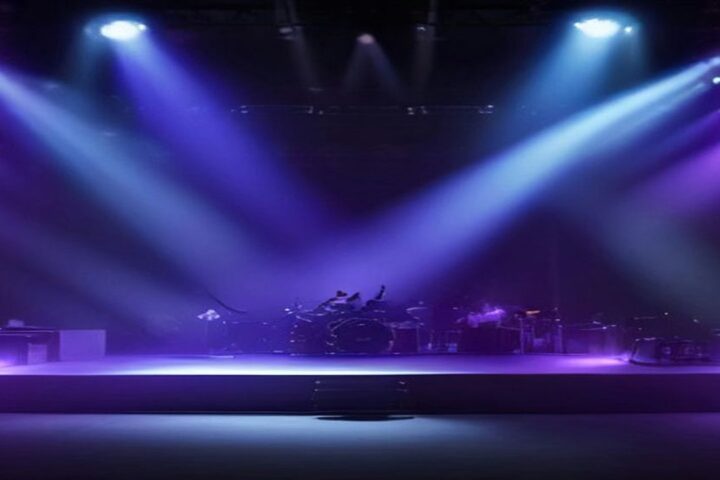 Escenario de concierto con batería, amplificadores y guitarras bajo haces de luz morada y azul que atraviesan la bruma