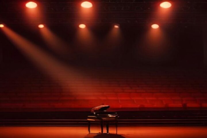 Escenario con un piano de cola iluminado por reflectores rojos y anaranjados frente a filas de butacas vacías