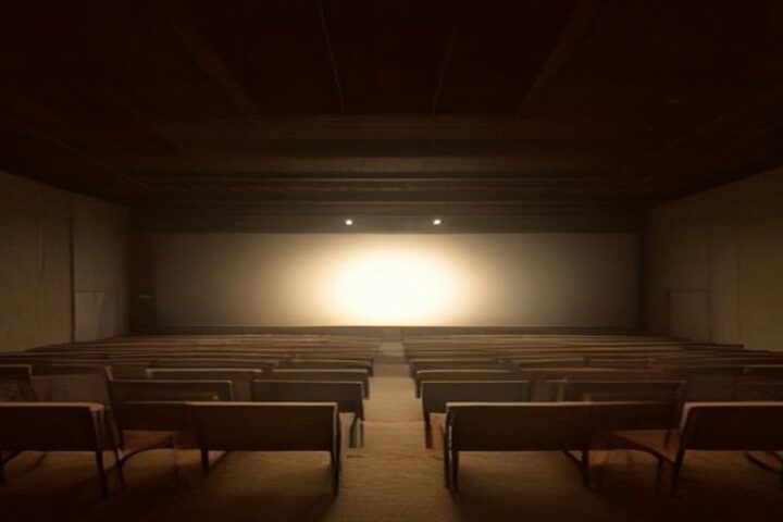Sala en penumbra con filas de bancas de madera vacías frente a un escenario iluminado por un haz de luz cálida