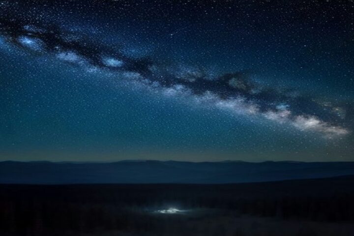 Cielo nocturno estrellado con la Vía Láctea sobre la silueta de un bosque y montañas, con un leve resplandor de ciudad en el horizonte