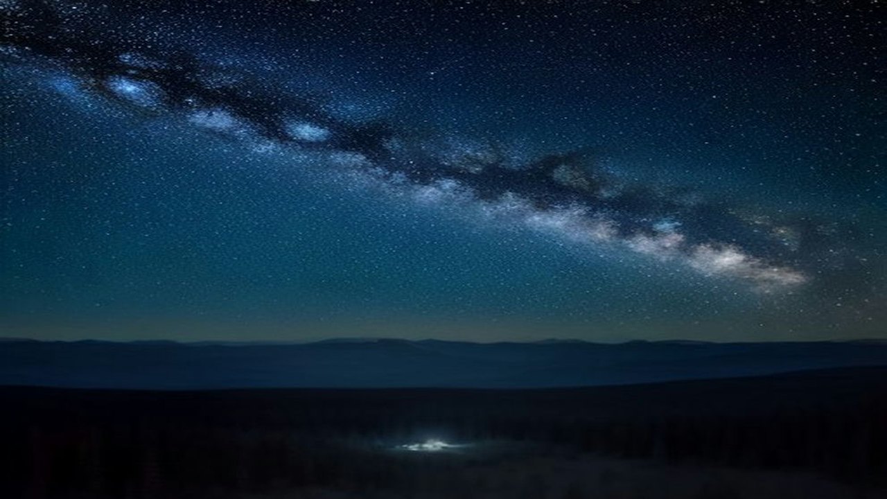 Cielo nocturno estrellado con la Vía Láctea sobre la silueta de un bosque y montañas, con un leve resplandor de ciudad en el horizonte