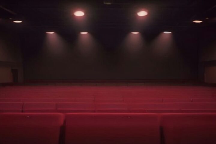 Sala de conciertos vacía con escenario oscuro iluminado por varios focos cálidos en el techo y filas de butacas rojas en primer plano