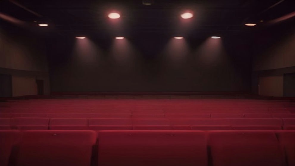 Sala de conciertos vacía con escenario oscuro iluminado por varios focos cálidos en el techo y filas de butacas rojas en primer plano