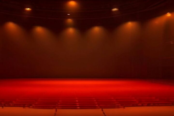 Sala de espectáculos vacía bañada en luz roja y anaranjada, con reflectores encendidos sobre un escenario amplio y sin público