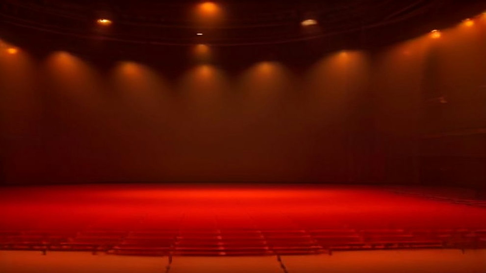 Sala de espectáculos vacía bañada en luz roja y anaranjada, con reflectores encendidos sobre un escenario amplio y sin público