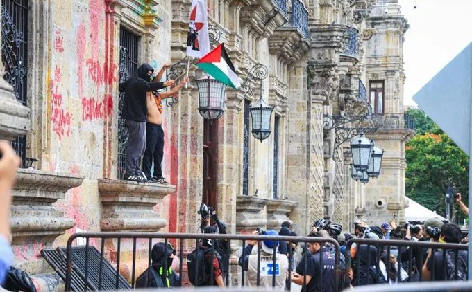 Personas encapuchadas cuelgan banderas, entre ellas una palestina, en un balcón de Palacio de Gobierno de Jalisco mientras abajo se concentran manifestantes y reporteros