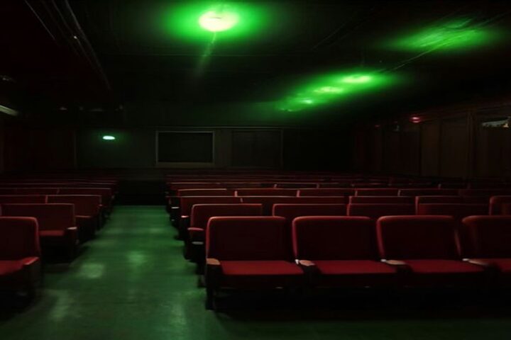 Sala de cine antigua con butacas rojas vacías, iluminada por destellos verdes en el techo y un ambiente de misterio