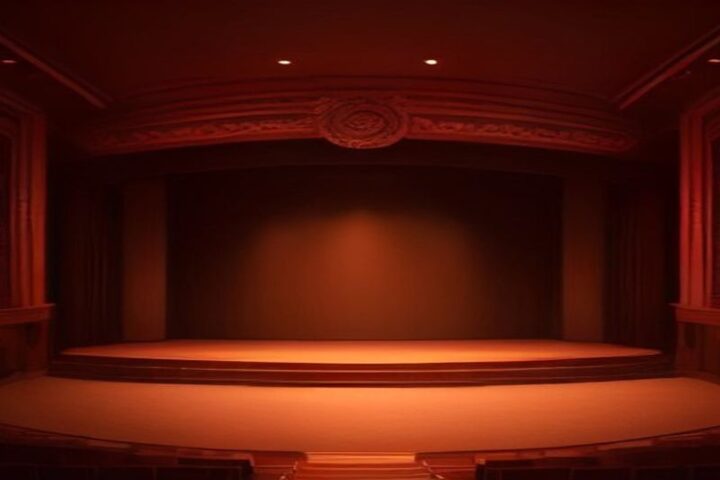 Escenario vacío de un teatro antiguo con arco ornamentado, telón oscuro y luz cálida rojiza sobre la tarima, visto desde las butacas en penumbra