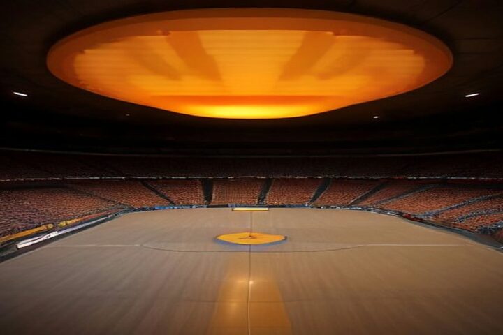 Arena techada vacía con una cancha de basquetbol de madera, gradas en penumbra y una gran lámpara anaranjada que ilumina el techo