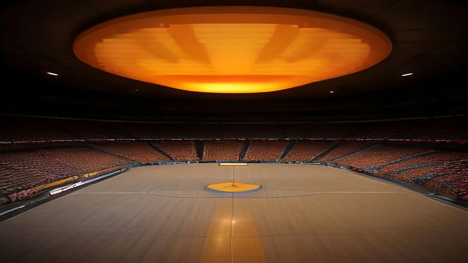 Arena techada vacía con una cancha de basquetbol de madera, gradas en penumbra y una gran lámpara anaranjada que ilumina el techo
