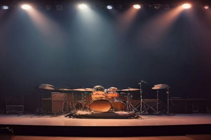 Escenario de concierto vacío con una batería al centro, amplificadores y micrófonos, iluminado por reflectores cálidos y azules frente a un telón oscuro