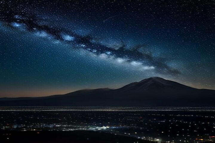 Cielo nocturno estrellado con la Vía Láctea y una estela de meteoro sobre la silueta de un volcán y las luces de una gran ciudad