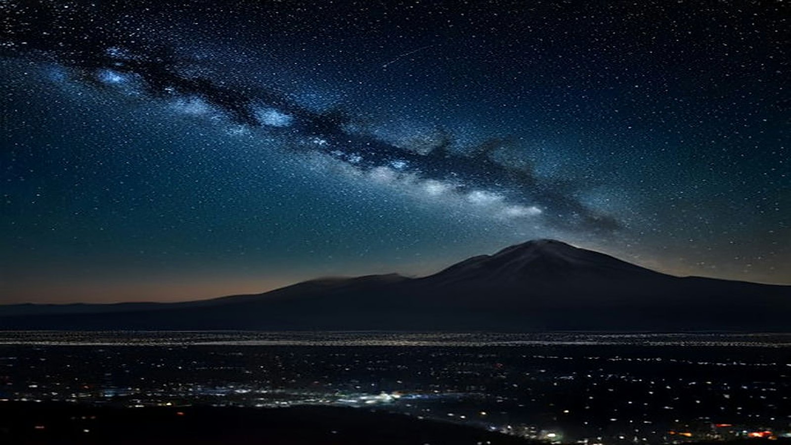 Cielo nocturno estrellado con la Vía Láctea y una estela de meteoro sobre la silueta de un volcán y las luces de una gran ciudad
