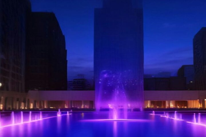 Plaza con fuentes iluminadas de violeta frente a edificios modernos de cristal al anochecer