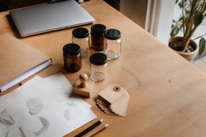 Mesa de madera de un taller emprendedor con frascos sin etiqueta, etiquetas kraft en blanco, sello de goma, bocetos a lápiz y una laptop cerrada