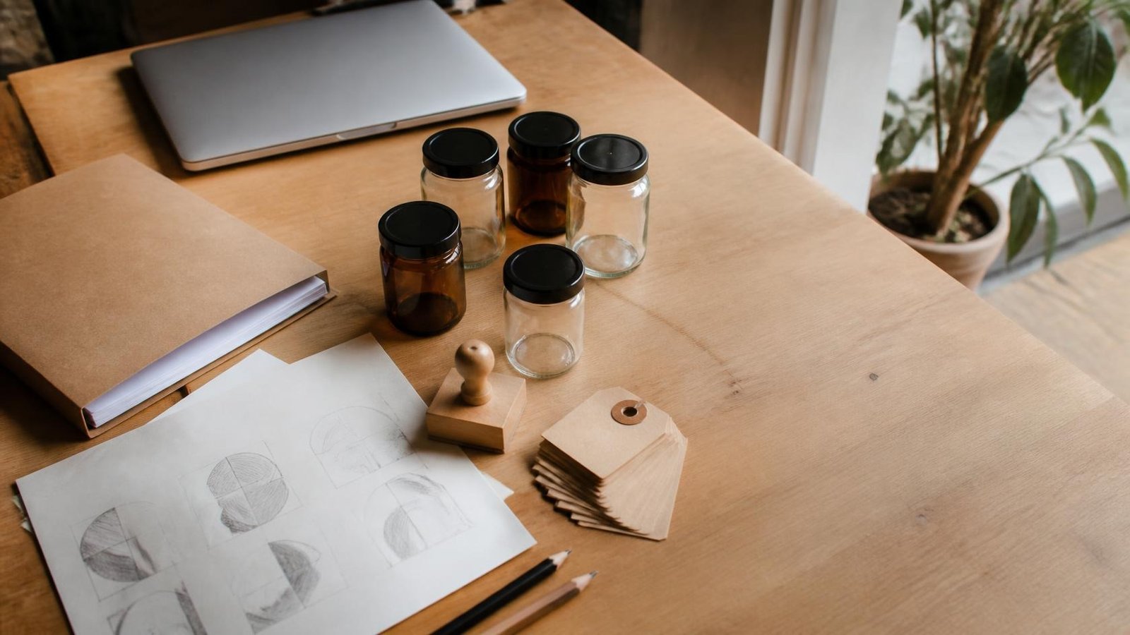 Mesa de madera de un taller emprendedor con frascos sin etiqueta, etiquetas kraft en blanco, sello de goma, bocetos a lápiz y una laptop cerrada