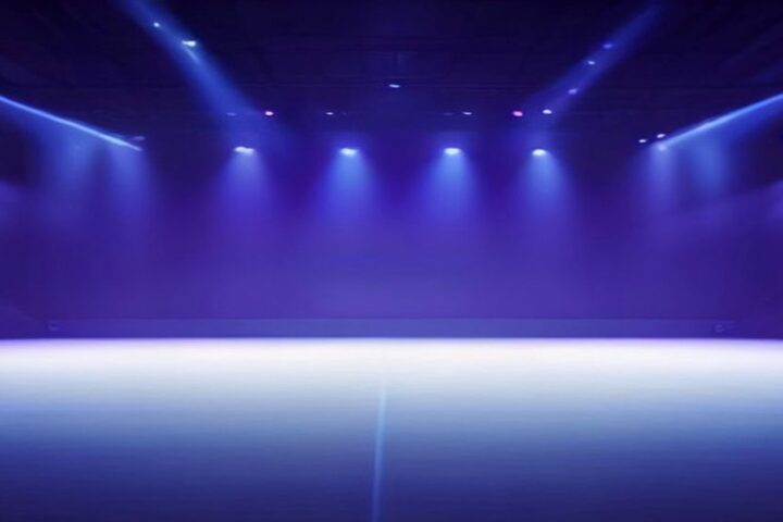 Escenario vacío de una arena de conciertos iluminado con reflectores azules y morados entre la bruma, sin público