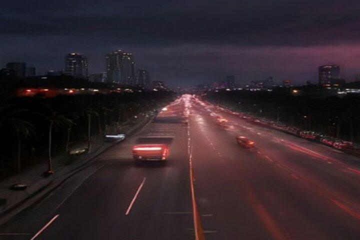 Avenida amplia de la Ciudad de México al anochecer vista desde un puente peatonal, con estelas de luces rojas de autos, pavimento húmedo, palmeras en el camellón y edificios iluminados bajo cielo nublado
