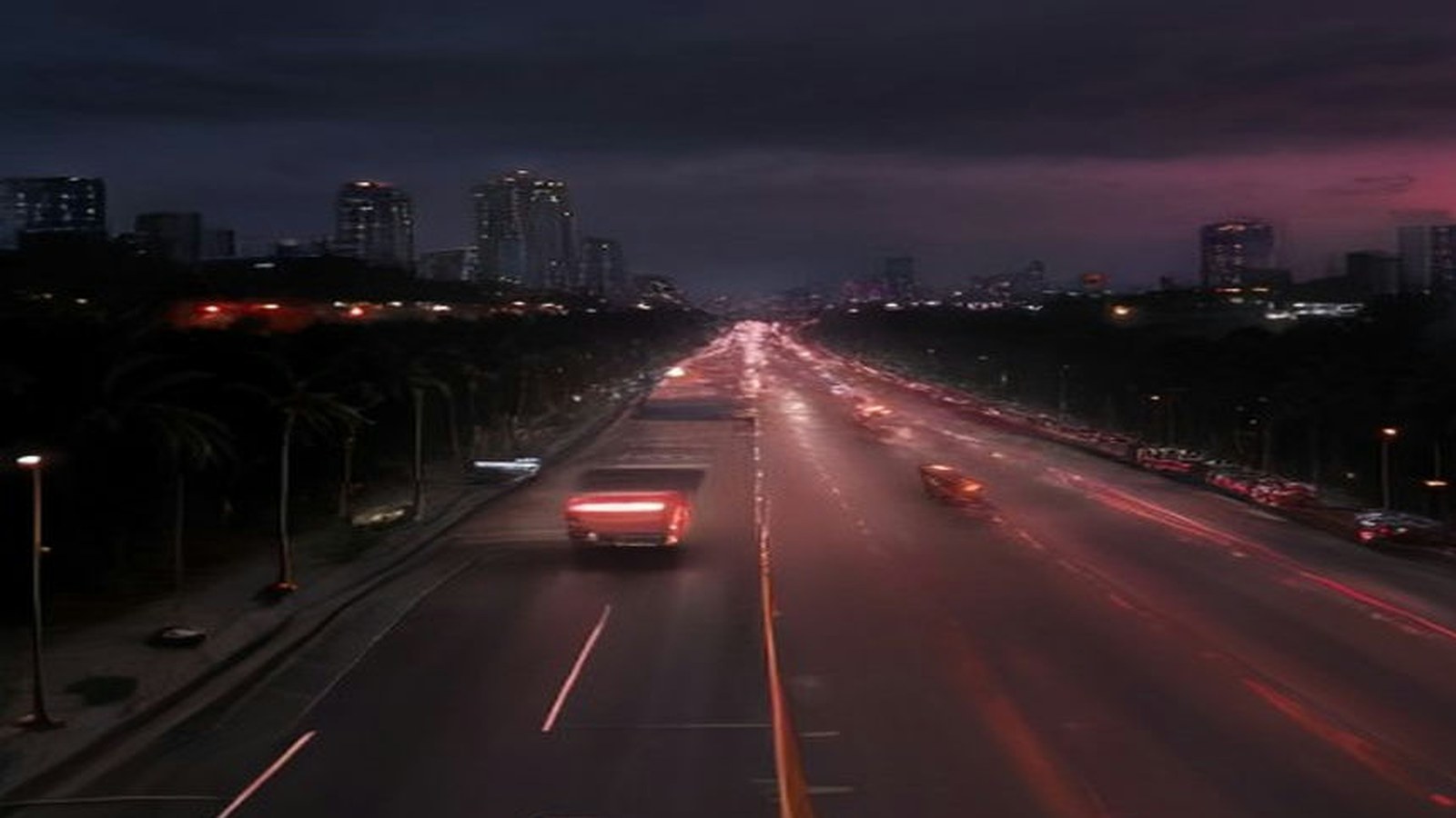 Avenida amplia de la Ciudad de México al anochecer vista desde un puente peatonal, con estelas de luces rojas de autos, pavimento húmedo, palmeras en el camellón y edificios iluminados bajo cielo nublado