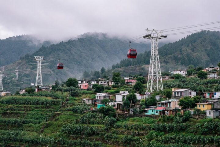 Cabinas de teleférico urbano suspendidas sobre campos de cultivo y casas de un pueblo en las laderas del sur de la Ciudad de México, con cerros verdes al fondo y cielo nublado