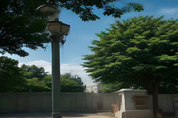Poste metálico con un equipo de cámara y antena en un parque arbolado de la ciudad bajo un cielo azul con nubes