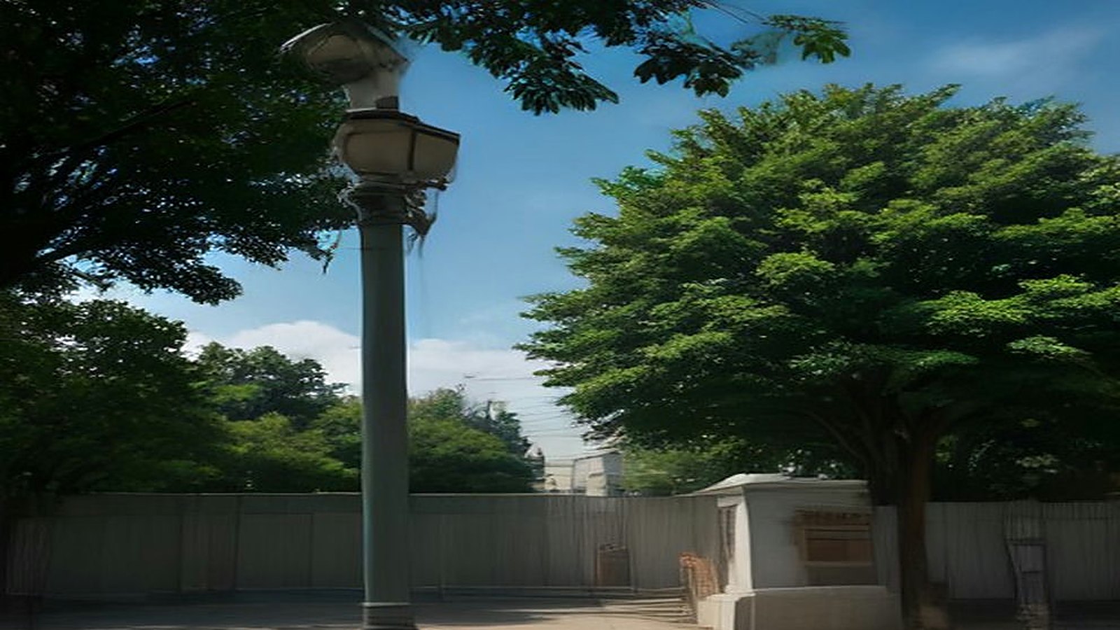 Poste metálico con un equipo de cámara y antena en un parque arbolado de la ciudad bajo un cielo azul con nubes
